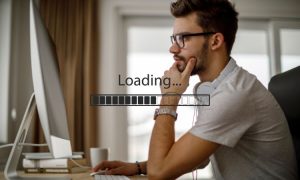 La imagen muestra a un hombre joven con barba y gafas sentado frente a una computadora de escritorio. Está concentrado, con la mano en la barbilla, en una postura pensativa. Lleva puestos unos auriculares blancos alrededor del cuello. En la pantalla de la computadora aparece una barra de carga con el texto "Loading..." (Cargando...). El ambiente parece ser una habitación luminosa con cortinas y ventanas al fondo.