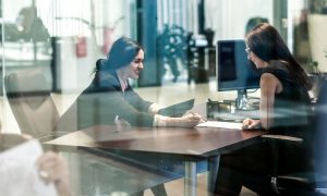 Imagen en la que se ven dos mujeres sentadas frente a frente en una oficina moderna. Ambas parecen estar en una reunión o entrevista de trabajo; una de ellas está escribiendo o tomando notas mientras la otra escucha atentamente. La escena transmite profesionalismo y colaboración en un entorno laboral. La fotografía está tomada a través de un vidrio, lo que añade un efecto de reflexión y profundidad.