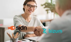 Una mujer joven, con gafas y una expresión sonriente, estrechando la mano de otra persona en un entorno de oficina. Sobre la imagen aparecen los logotipos de SIDN Digital Thinking y Bizum, simbolizando un acuerdo o alianza entre ambas compañías. La escena transmite profesionalidad, confianza y colaboración.