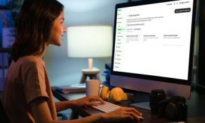 Persona usando un ordenador, sentada frente a una pantalla grande en una oficina con iluminación suave. Té o café y cámara en la mesa. Marketing digital.