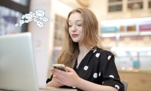 imagen de una mujer joven sentada frente a un portátil, mientras sostiene un teléfono móvil. Viste una blusa negra con lunares blancos y parece concentrada en su actividad digital. En la parte superior izquierda de la imagen, se visualiza un gráfico ilustrativo de sobres flotando, que simboliza el envío de correos electrónicos, probablemente relacionado con email marketing o comunicación digital. El entorno sugiere un espacio moderno y luminoso, como una cafetería o una oficina creativa.