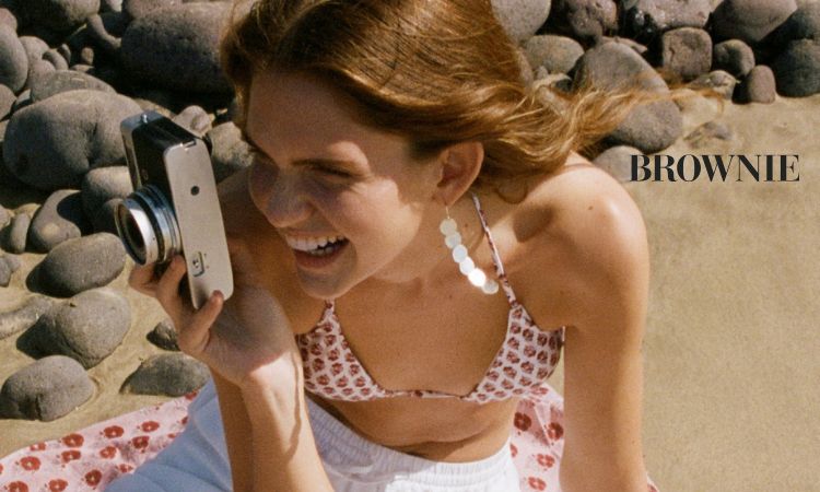 Mujer sonriente sostiene cámara, sentada en playa rocosa. Texto: "BROWNIE". Contexto veraniego, moda y estilo.