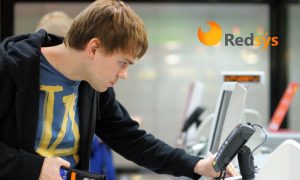 Fotografía de un hombre de unos 25 años utilizando una TPV para pagar. El hombre mira la TPV con extrañeza, representando la caída de Redsys