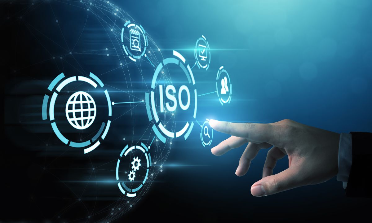 una mano humana tocando una interfaz digital con iconos tecnológicos flotantes. En el centro se destaca el texto “ISO”, representando los estándares internacionales de calidad y gestión. A su alrededor aparecen símbolos relacionados con la conectividad global, la configuración, el calendario y la gestión de usuarios, todo sobre un fondo azul futurista que evoca innovación y certificación tecnológica.