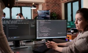 Imagen de dos mujeres charlando, con cara de preocupación, frente a un PC con códigos y html en la pantalla.