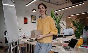 Persona sonriente sostiene cajas, de pie en oficina moderna con pizarra blanca, plantas y portátil. Entorno dinámico, enfoque en e-commerce y logística.