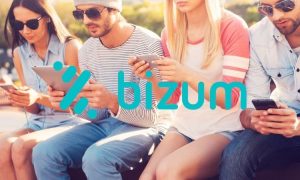 Fotografía de un grupo de mujeres y hombres de entre unos 20 y 35 años sentados al aire libre mientras usan sus teléfonos. En el centro, sobre la imagen, aparece el logo de Bizum