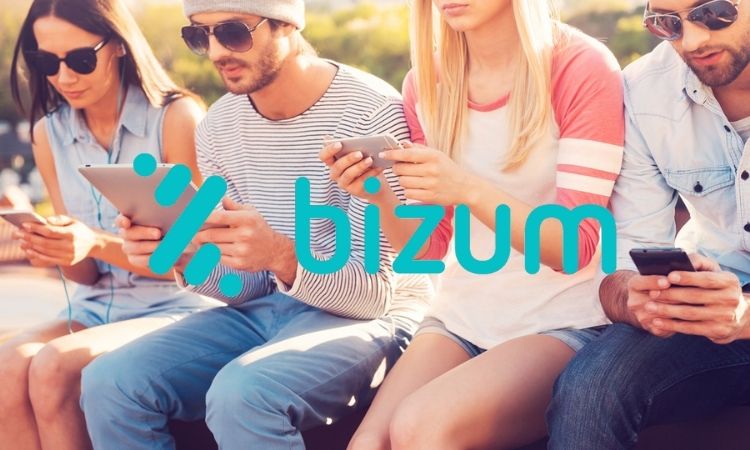 Fotografía de un grupo de mujeres y hombres de entre unos 20 y 35 años sentados al aire libre mientras usan sus teléfonos. En el centro, sobre la imagen, aparece el logo de Bizum