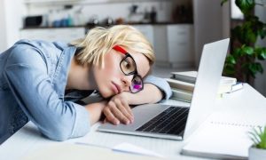 Una persona, apoyada sobre una mesa, mira con cansancio su portátil en una oficina, rodeada de libros, reflejando agotamiento laboral.