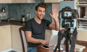 Un hombre sonríe y saluda a una cámara mientras escribe en un portátil. Se encuentra en una cocina moderna. Ideal para marketing digital.