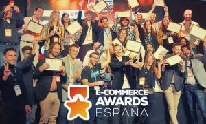 Grupo de personas sonrientes sosteniendo certificados de premiación sobre un escenario. Texto visible: "E-COMMERCE AWARDS ESPAÑA". Ambiente festivo y profesional.