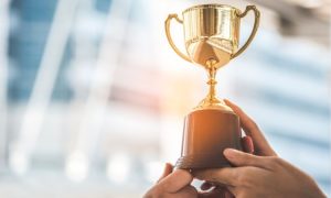 Trofeo dorado sostenido por manos; representa éxito y logro. Brilla bajo la luz en un ambiente urbano desenfocado. Texto ausente.