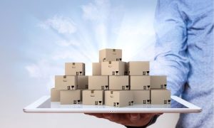 Cajas apiladas sobre una tablet sostenida por una mano; fondo de cielo despejado. Representa logística y gestión de inventarios digitales.