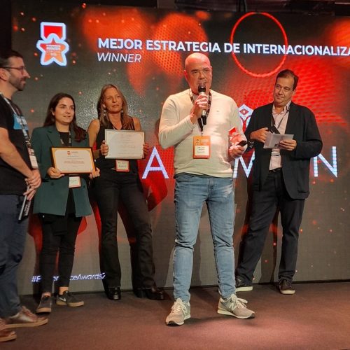 Persona habla en micrófono, rodeada de cuatro personas con certificados, en ceremonia de premiación. Texto: "MEJOR ESTRATEGIA DE INTERNACIONALIZACIÓN WINNER".