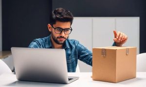 un hombre joven en actitud pensativa. Está sentado ante un ordenador portátil que está sobre una mesa. En una mano, el hombre sostiene una caja de cartón cerrada y precintada