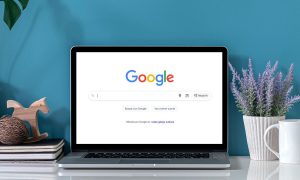 Fotografía de un ordenador portátil colocado sobre un escritorio con plantas y libretas ordenadas. En la pantalla del ordenador se ve la vista de inicio del buscador de Google