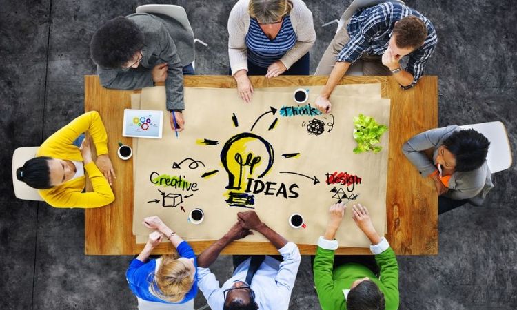 Personas trabajando juntas alrededor de una mesa con un boceto de bombilla y la palabra "IDEAS." Contexto de colaboración creativa y lluvia de ideas.