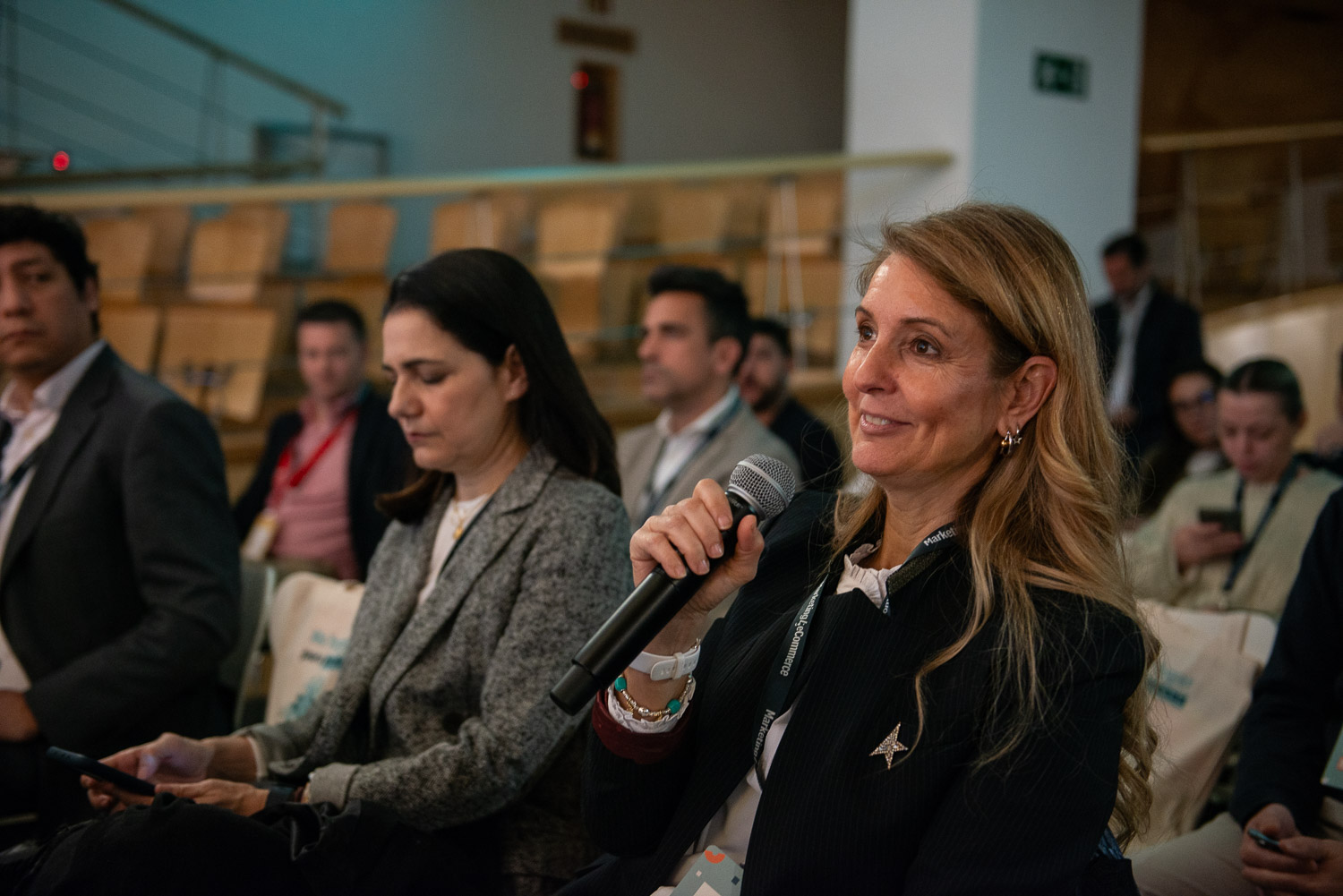 Mujer con micrófono sonríe; está en una conferencia. Varias personas sentadas escuchando. Texto visible: “Marketplace”. Ambiente profesional y de networking.