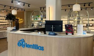 Mostrador moderno en tienda óptica; pantalla en el centro, estanterías iluminadas detrás con productos. Texto visible: "e-lentillas". Ambiente acogedor de óptica.