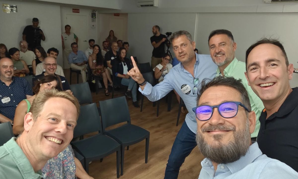 Aquí los cinco ponentes toman un selfie desde el frente de la sala, con el público asistente al fondo, sonriendo y participando. En la audiencia se percibe interés y complicidad, evidenciando el éxito del formato dinámico, en el que las preguntas del moderador generaron comparaciones entre plataformas y aportaron un toque divertido al debate.