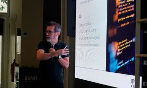 Un ponente, micrófono en mano, explica un concepto señalando una pantalla grande con texto y código. La sala es interior, con luz tenue, y el orador aparece concentrado en su exposición.