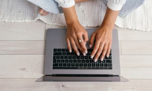 Manos tecleando en un portátil sobre un suelo claro; contexto de trabajo desde casa, representación de productividad y tecnología.