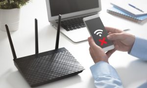 Fotografía de una mesa en la que están colocados un router y un ordenador portátil. Una persona tiene sus brazos apoyados en la mesa mientras sostiene un teléfono móvil cuya pantalla indica que no hay señal ni Internet.