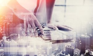 Fotografía de las manos de una mujer utilizando una tablet en la que se muestran gráficas de resultados. A modo de transparencia, sobre esa imagen se ha aplicado otra fotografía de una ciudad repleta de altos edificios.
