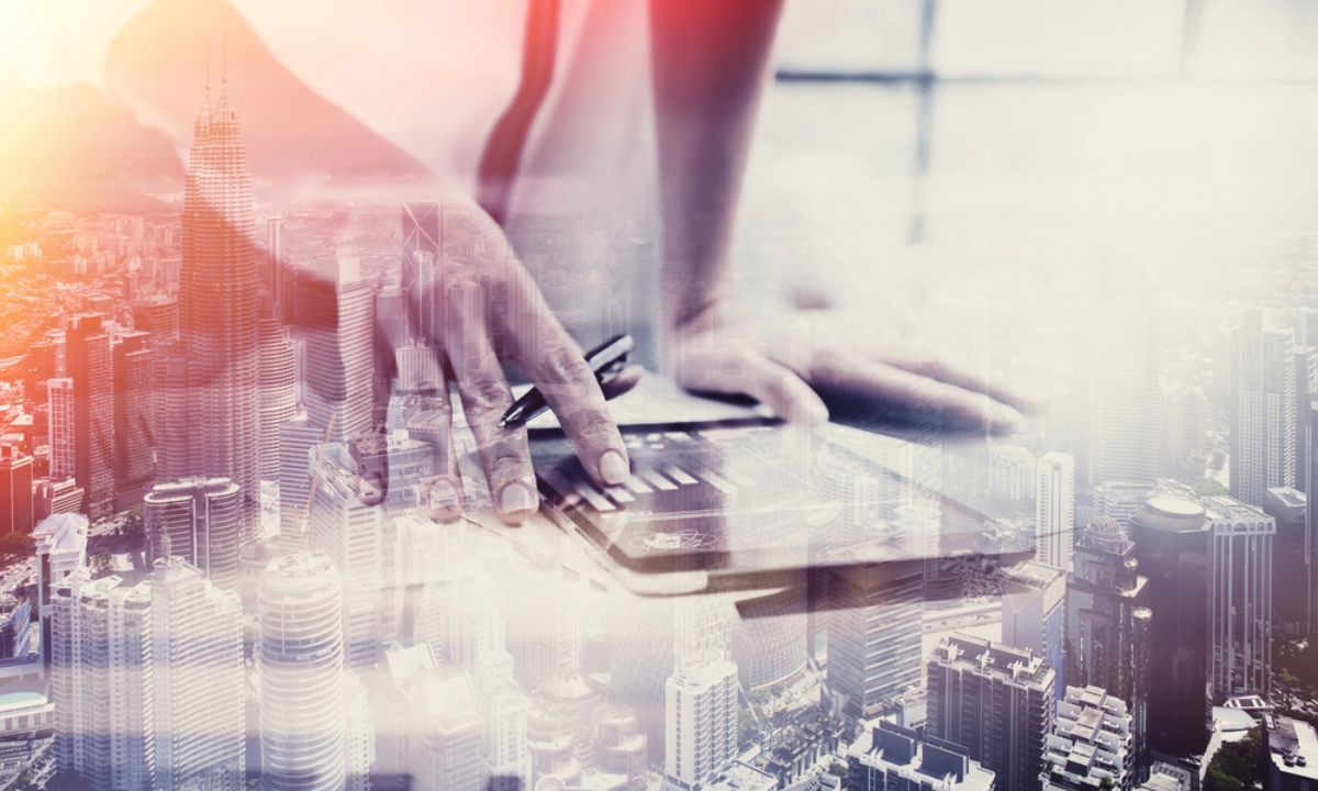 Fotografía de las manos de una mujer utilizando una tablet en la que se muestran gráficas de resultados. A modo de transparencia, sobre esa imagen se ha aplicado otra fotografía de una ciudad repleta de altos edificios.