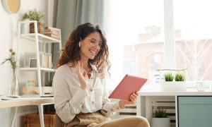 La imagen muestra a una mujer joven, con cabello rizado y gafas, sonriendo mientras sostiene una tablet de color rosa y saluda. Ella está sentada en un ambiente luminoso y moderno, con estanterías a su alrededor y plantas decorativas. Está usando unos auriculares inalámbricos, y parece estar en una videollamada o interactuando con la tablet de manera amigable.