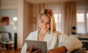 La imagen muestra a una mujer joven de cabello rubio, sonriendo mientras sostiene una tablet y la observa con atención. Está en un entorno acogedor, posiblemente su hogar, con luz cálida y mobiliario moderno al fondo.