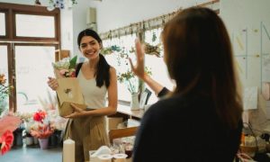 Dos mujeres despidiendose en una floristería. Una de ellas es una clienta, que lleva una planta empaquetada en brazos, y la otra es la dependienta. Se despiden de forma amigable, haciendo alusión al tema del artículo: la fidelización de clientes.