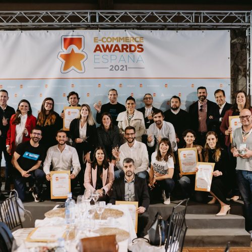 Grupo de personas posando con certificados frente a un cartel que dice "ECOMMERCE AWARDS 2021". Contexto de evento con mesas y sillas alrededor.