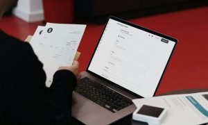 La imagen muestra a una persona gestionando facturas. En una mano sostiene una factura impresa, mientras en la otra utiliza un ordenador portátil (MacBook) con una interfaz de software de facturación electrónica abierta. En la mesa también hay documentos y un lector de tarjetas o dispositivo electrónico pequeño. El entorno sugiere un espacio de trabajo moderno, con un suelo rojo y mobiliario minimalista.