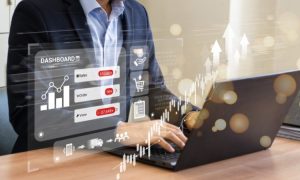 Persona usando ordenador portátil; expresa analíticas de datos: barra, gráfico de flechas ascendentes y texto: "DASHBOARD", "Metric", "Retail", "Service"; en oficina.