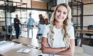 Persona sonriente, de pie con los brazos cruzados, en una oficina moderna con personas trabajando al fondo. Contexto de colaboración y éxito.