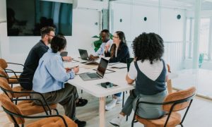 Fotografía de un grupo de profesionales compuesto por tres mujeres y dos hombres de etnias diversas que están sentados en el interior de una sala de oficina trabajando en un proyecto de inversión publicitaria.