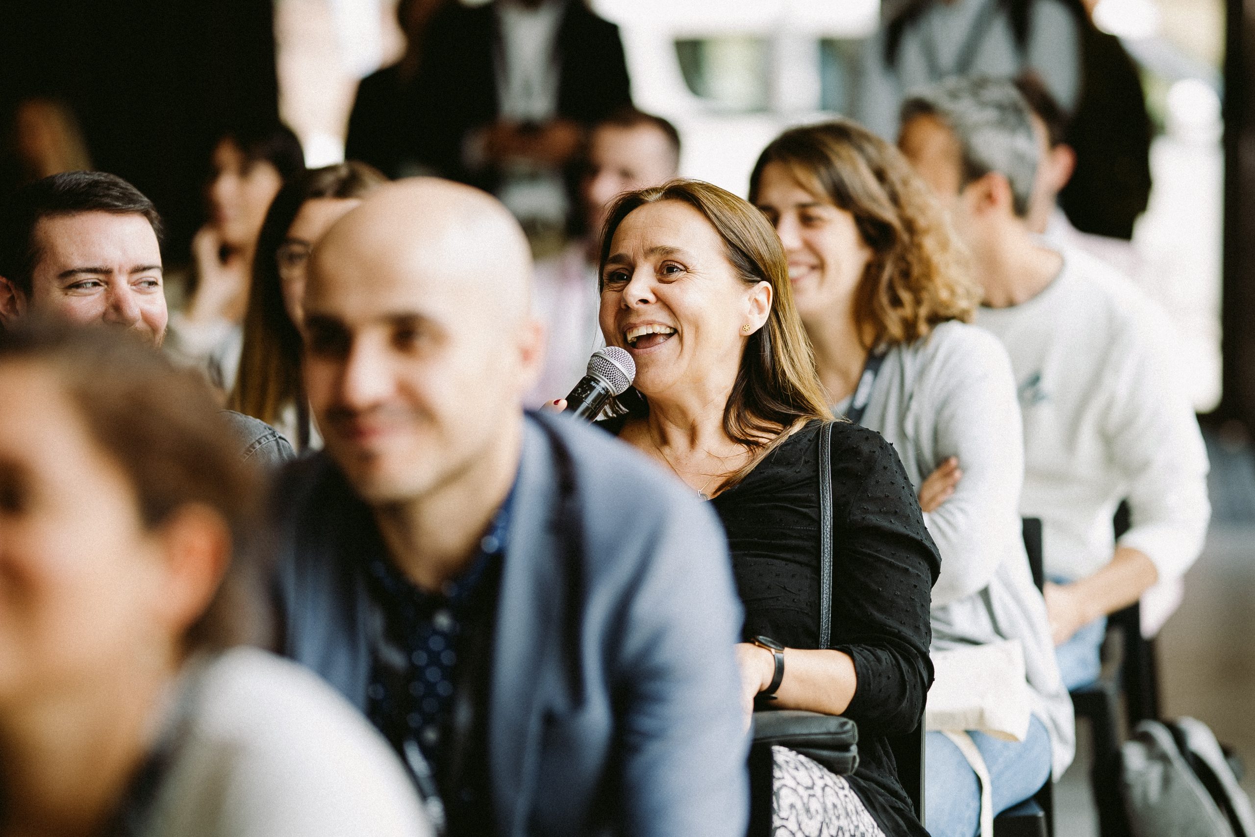 Persona hablando al micrófono, sonriendo, en una conferencia; asistentes alrededor atentos y un fondo difuminado. Ambiente de networking y participación.