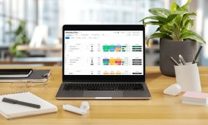Portátil mostrando un panel de control de marketing digital con gráficos y datos. Se sitúa sobre un escritorio con plantas, cuaderno y ratón.