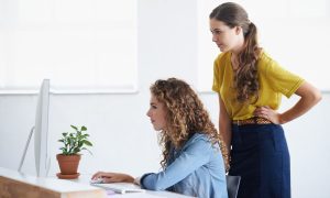 La imagen muestra a dos mujeres jóvenes en una oficina, concentradas frente a un ordenador de sobremesa. Una de ellas, con el cabello rizado y suelto, está sentada escribiendo en el teclado, mientras la otra, de cabello largo recogido en una coleta, permanece de pie a su lado observando la pantalla con atención. Sobre el escritorio hay una pequeña maceta con una planta. El entorno es luminoso y minimalista, con paredes blancas y ventanas grandes al fondo.