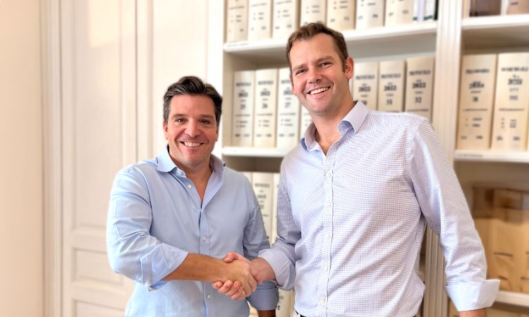 Dos hombres estrechan manos, sonriendo, en una oficina con estanterías llenas de carpetas. Acuerdo empresarial, colaboración exitosa y ambiente profesional.