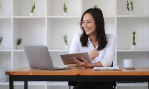 Una mujer joven, con gafas y camisa blanca, sentada en un escritorio de madera frente a un portátil abierto. Ella sonríe mientras sostiene una tablet con una mano y un bolígrafo con la otra, en un ambiente moderno y ordenado con estanterías blancas al fondo decoradas con pequeñas plantas. La escena transmite profesionalidad y un ambiente relajado, ideal para ilustrar conceptos de trabajo digital, marketing de contenidos o formación online.