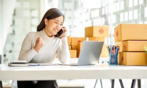 Una mujer sonriente en una oficina luminosa, hablando por teléfono mientras trabaja en un portátil. Está rodeada de cajas de cartón, lo que sugiere que gestiona un negocio de comercio electrónico o logística. Su gesto con el puño cerrado indica entusiasmo o éxito, posiblemente por una buena noticia relacionada con su actividad profesional.