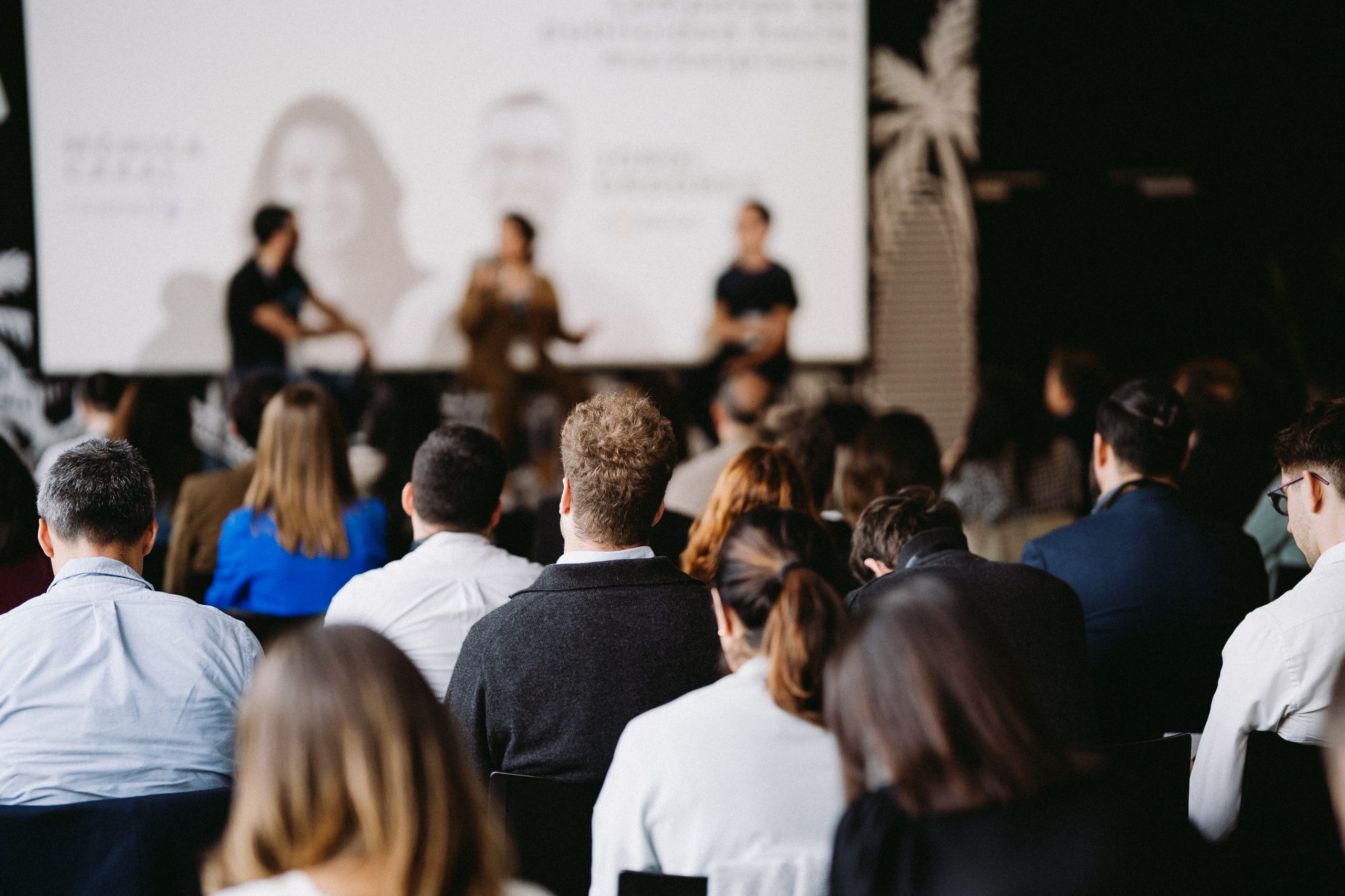 Personas sentadas, escuchando una conferencia; al frente, oradores hablando, proyector de fondo. Ambiente profesional y de networking.