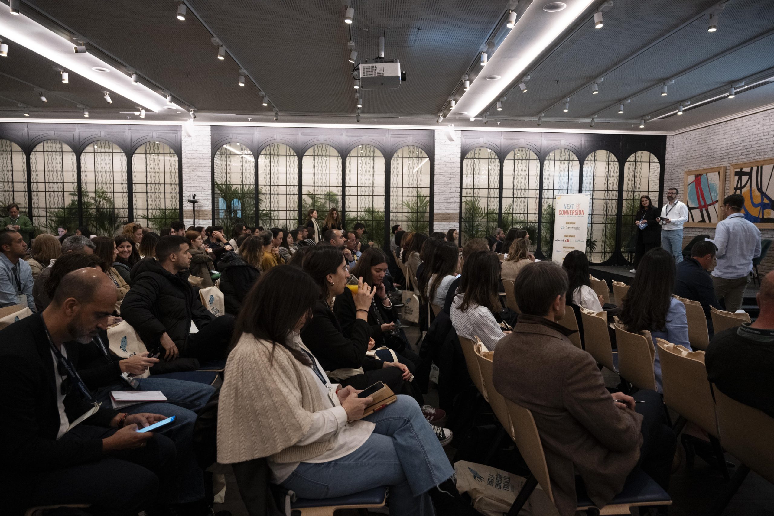 Personas sentadas atendiendo una presentación en un salón moderno con carteles. Texto visible: "NEXT CONVERSION 2023". Ambiente profesional. Marketing.