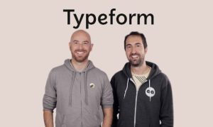 Dos personas de pie, sonriendo, con sudaderas grises; fondo beige. Texto: "Typeform". Ideal para contenido de marketing y colaboración dinámica.