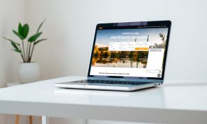 Portátil mostrando una página web de viajes con texto: "Encuentra hoteles. Protegido con Paycomet". Está sobre una mesa blanca, planta en el fondo. Ambiente moderno.