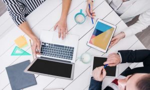 Portátil y tablet en mesa blanca; manos trabajando, resaltadores y gráficos coloridos; entorno de oficina; colaboración y estrategia.