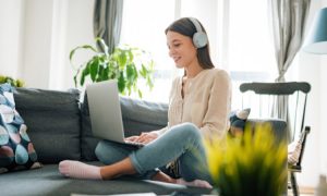 Una joven sentada en un sofá, con auriculares puestos, trabajando o disfrutando de algo en su ordenador reacondicionado. Ella está en un ambiente luminoso y cómodo, con plantas y decoraciones en el fondo. La mujer lleva una blusa clara y jeans, y se ve relajada mientras usa su dispositivo.