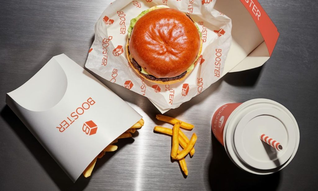 Fotografía en plano cenital de un menú de comida rápida compuesto por una hamburguesa, una caja de patatas y un refresco. Todos los elementos lucen un packaging personalizado con el logo de Booster